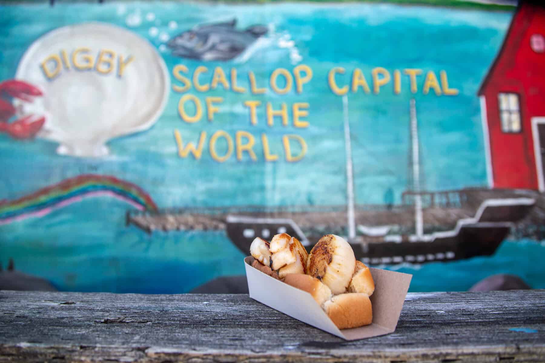 A scallop roll in front of the Scallop Capital of the World Mural in Digby, Nova Scotia